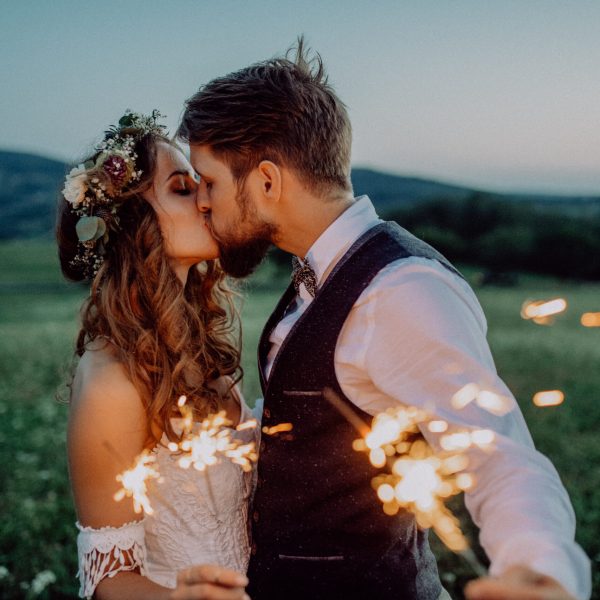 Beautiful young bride and groom on a meadow in the evening, holding sparklers. A man and a woman kissing.