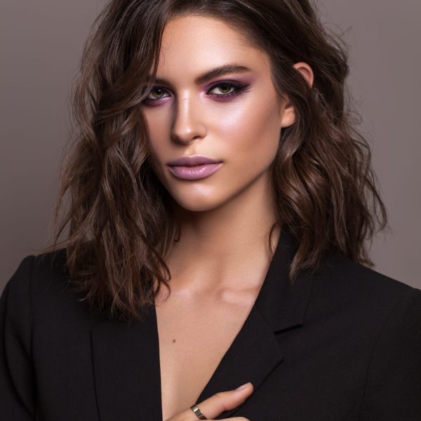Portrait of young beautiful brunette model with violet professional make up, trendy wavy hairdo and perfect skin. Confident business lady in black fashion jacket on a grey background.